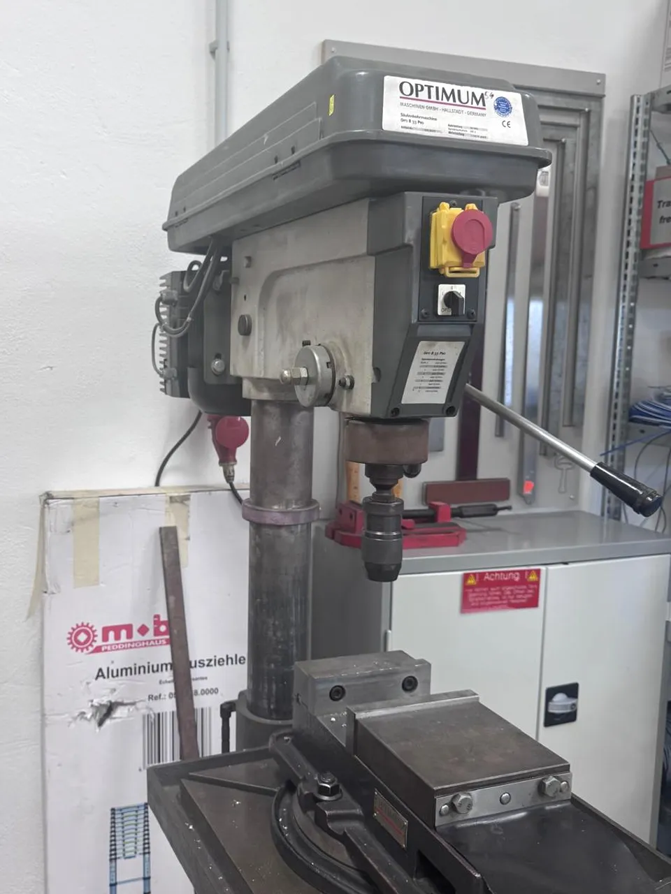 Säulenbohrmaschine in einem gewerblichen Arbeitsbereich, mit Schaltkasten und rotem Schalter am Maschinenkopf.